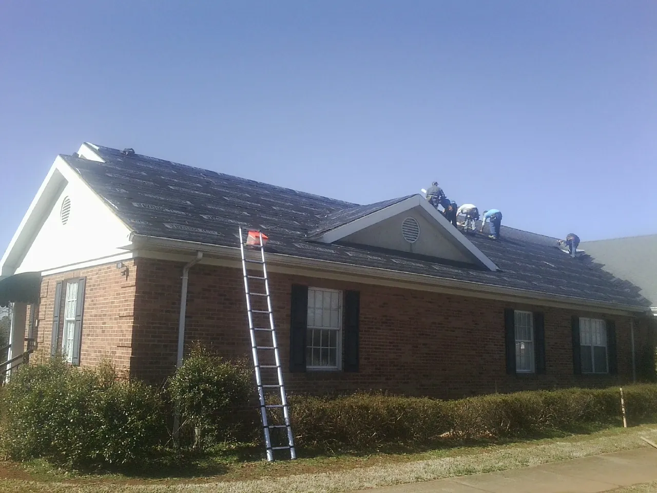 Crew tearing shingles off a brick ranch roof with a ladder set — Metal Roof Repair work by our crew in Fort Jones