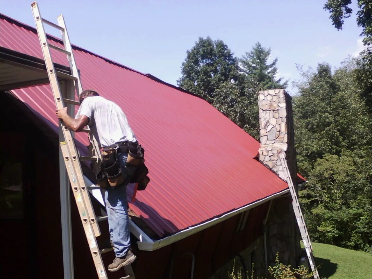 Roofer on a ladder beside a red metal roof and stone chimney — Soffit & Fascia Repair by Roofing Brothers in Fort Jones